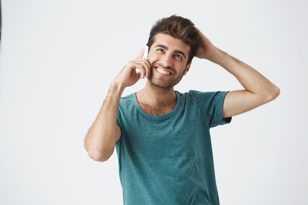 Ritratto di giovane maschio ispanico attraente che sorride in camicia blu, con la bella acconciatura, essendo timido mentre parla al telefono con la sua ragazza.