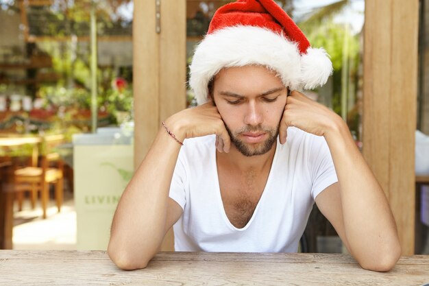 Ritratto di giovane maschio sconvolto vestito con maglietta bianca e cappello di Babbo Natale rosso sensazione di malessere dopo aver bevuto con gli amici alla festa di Capodanno