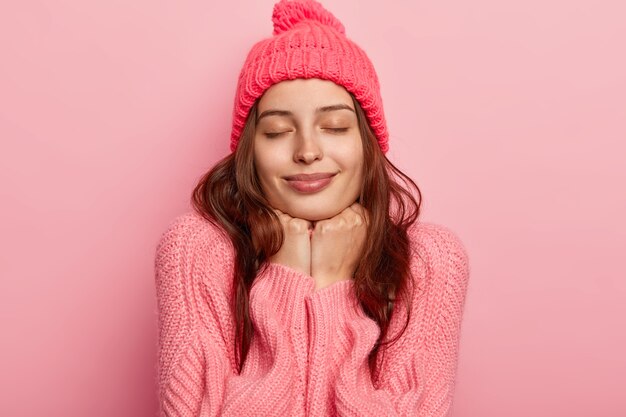 Ritratto di giovane modello femminile rilassato tiene entrambe le mani sotto il mento, ha gli occhi chiusi, indossa un cappello e un maglione rosei, si sente soddisfatto, posa su sfondo rosa.