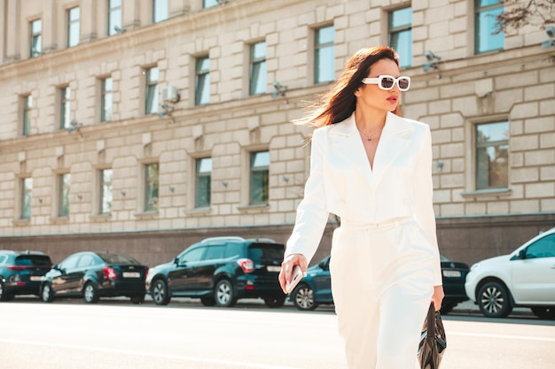 Ritratto di giovane modello moderno sexy della donna di affari. Bella donna calda in abito bianco in posa sullo sfondo della strada. Moda femminile che cammina all'aperto in occhiali da sole