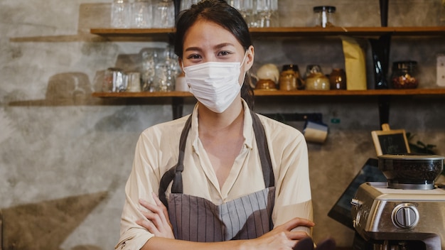 Ritratto di giovane ragazza asiatica cameriera indossare maschera medica sensazione di sorriso felice in attesa di clienti dopo il blocco al caffè urbano.