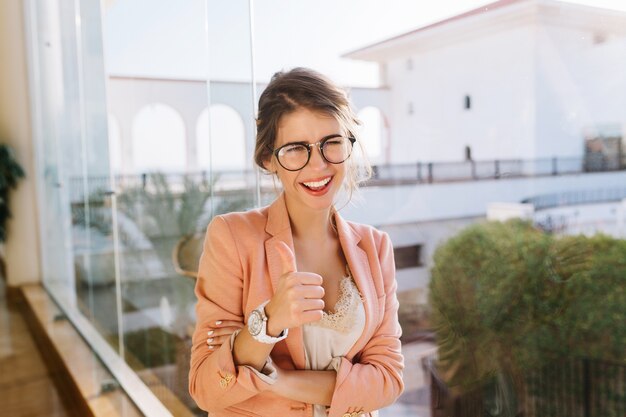 Ritratto di giovane ragazza che indossa occhiali alla moda, signora intelligente in elegante giacca rosa con camicetta beige, studente felice che mostra i pollici in su. Grande finestra con bella vista.