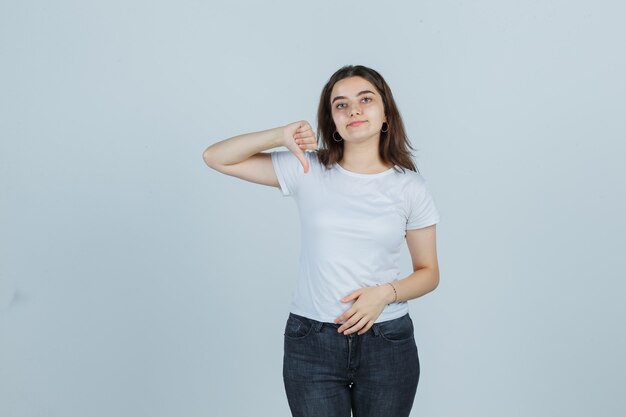 Ritratto di giovane ragazza che mostra il pollice verso il basso in t-shirt, jeans e guardando fiducioso vista frontale