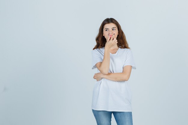 Ritratto di giovane ragazza che pizzica le labbra in t-shirt bianca, jeans e guardando premurosa vista frontale