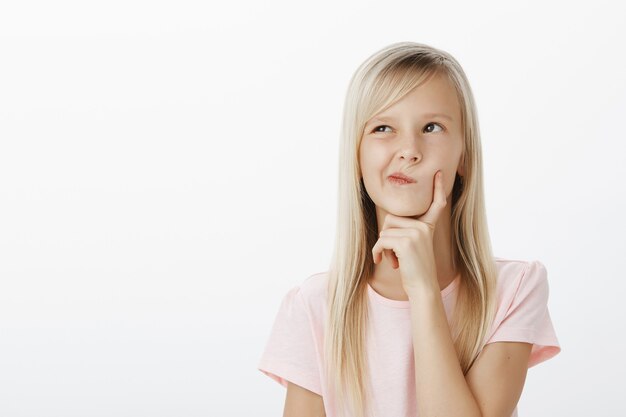 Ritratto di giovane ragazza creativa premurosa con capelli biondi lunghi, alzando lo sguardo e tenendo la mano sul mento mentre pensa o calcola in mente