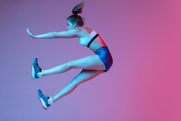 Ritratto di giovane ragazza sportiva in allenamento sportivo che salta isolato su sfondo rosa studio in luce al neon