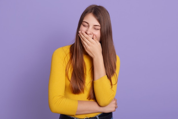 Ritratto di giovane ragazza stanca che indossa un maglione giallo contro il muro lilla, coprendosi la bocca con le palme