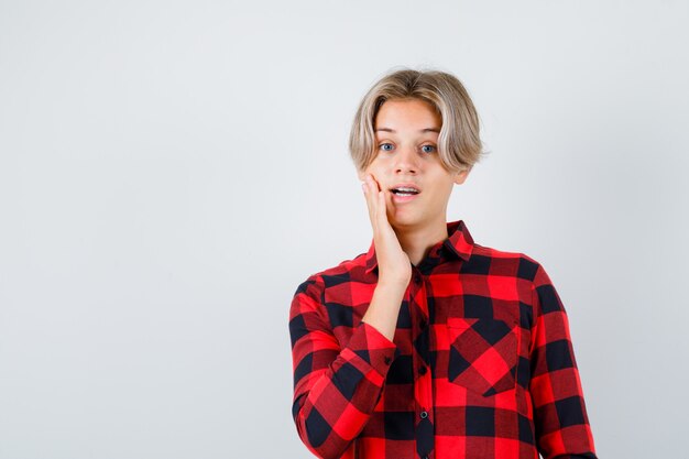 Ritratto di giovane ragazzo adolescente con la mano sulla guancia in camicia a quadri e guardando sorpreso vista frontale