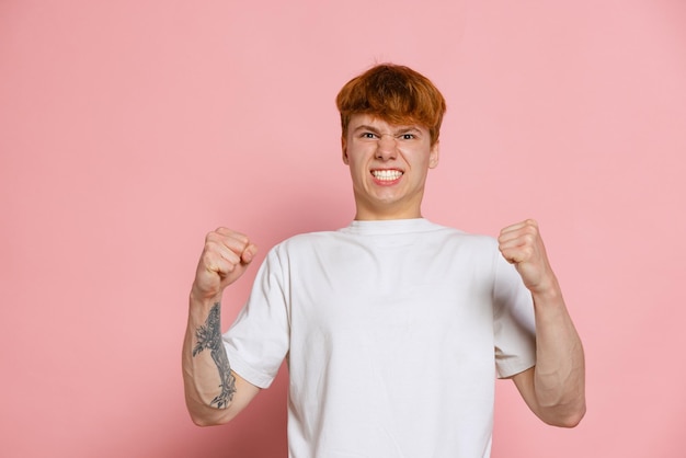 Ritratto di giovane ragazzo dai capelli rossi in panno casual in posa isolato su sfondo rosa studio Pugni in su