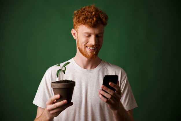 Ritratto di giovane sorridente bello rosso barbuto giovane, tenendo la pianta maculata, guardando il telefono cellulare