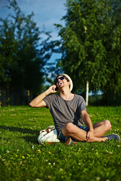 Ritratto di giovane sorridente ridendo attraente elegante uomo moderno in panno casual nel cappello con gli occhiali seduto nel parco in erba verde parlando al telefono