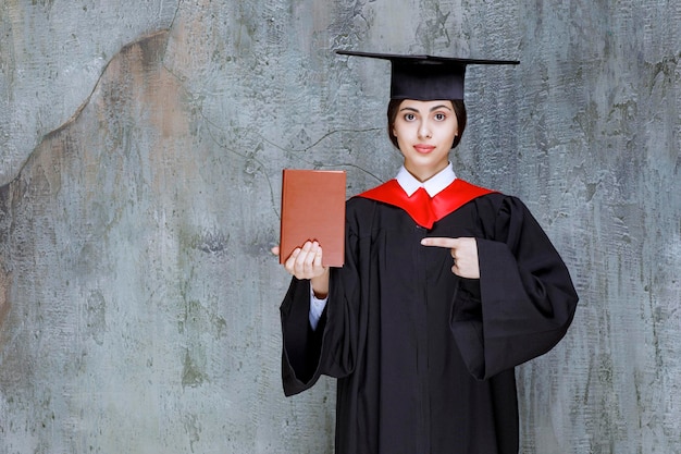 Ritratto di giovane studente laureato con libro in piedi contro il muro. Foto di alta qualità
