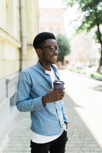 Ritratto di giovane uomo africano felice che cammina sulla strada con la tazza di caffè.