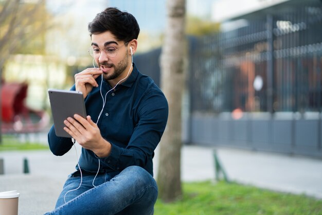 Ritratto di giovane uomo avente una videochiamata sulla tavoletta digitale mentre è seduto su una panchina all'aperto. Concetto urbano.