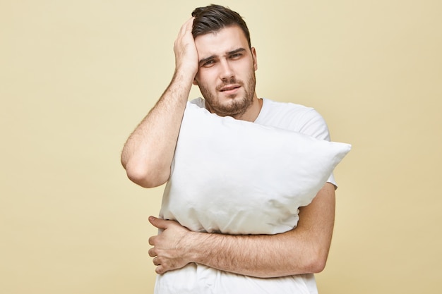 Ritratto di giovane uomo castano stressato che soffre di mal di testa tenendo la mano sulla testa e tenendo il cuscino non può addormentarsi senza sonniferi, avendo depresso l'espressione facciale frustrata