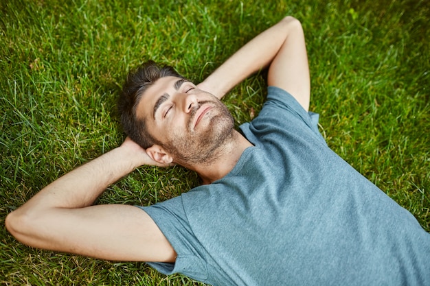 Ritratto di giovane uomo caucasico di bell'aspetto maturo in camicia blu pacifica sdraiato sull'erba con sì chiuso.