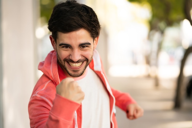 Ritratto di giovane uomo che celebra la vittoria stando in piedi all'aperto in strada. Concetto urbano e di successo.