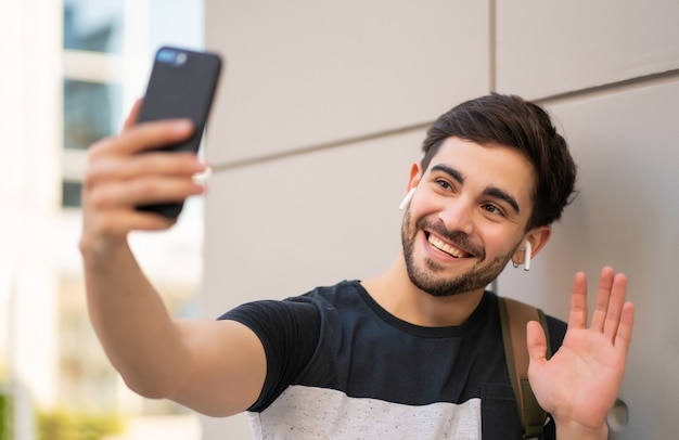 Ritratto di giovane uomo che ha una videochiamata sul telefono cellulare mentre si sta in piedi all'aperto. Concetto urbano.