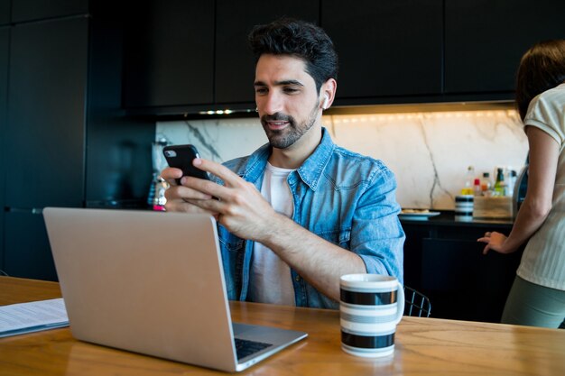 Ritratto di giovane uomo che lavora con un computer portatile e un telefono cellulare da casa mentre la donna cucina in background