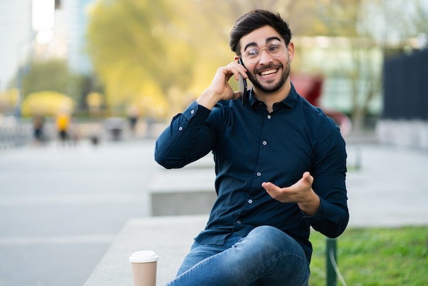 Ritratto di giovane uomo che parla al telefono mentre è seduto su una panchina all'aperto. Concetto urbano.