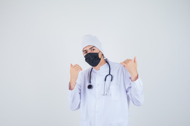 Ritratto di giovane uomo che punta a se stesso in uniforme bianca, maschera e guardando fiducioso vista frontale