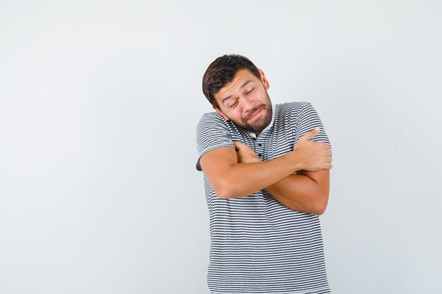 Ritratto di giovane uomo che si abbraccia, tenendo gli occhi chiusi in maglietta e guardando mite vista frontale