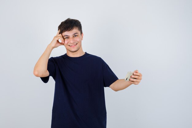 Ritratto di giovane uomo che tiene il telefono cellulare, in piedi nella posa di pensiero in maglietta nera e guardando allegro vista frontale