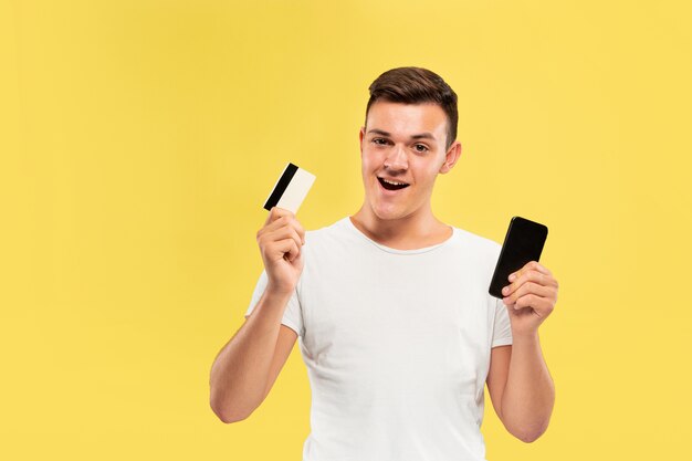 Ritratto di giovane uomo che tiene smartphone e carta di credito isolato sulla parete gialla