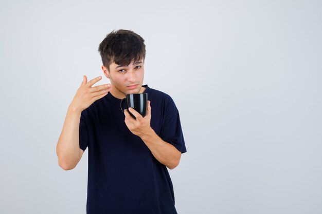 Ritratto di giovane uomo che tiene tazza di tè, indicando lontano in maglietta nera e guardando pensieroso vista frontale