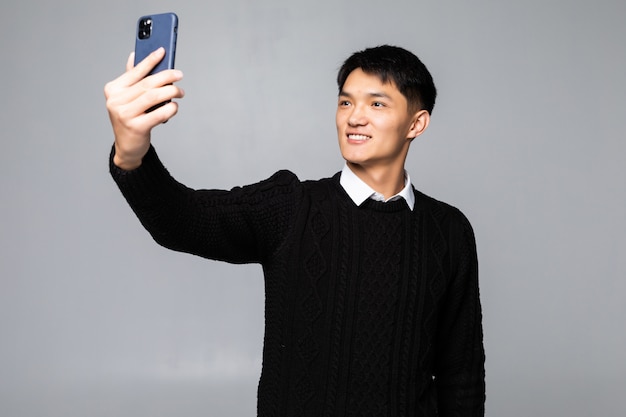 Ritratto di giovane uomo cinese sorridente che prende un selfie con il telefono cellulare mentre isolato sopra la parete bianca