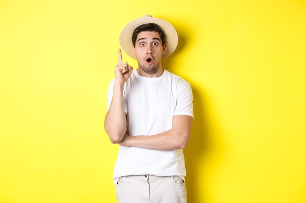 Ritratto di giovane uomo con cappello di paglia che ha un'idea, alzando il dito segno eureka, dando suggerimenti, in piedi su sfondo giallo.