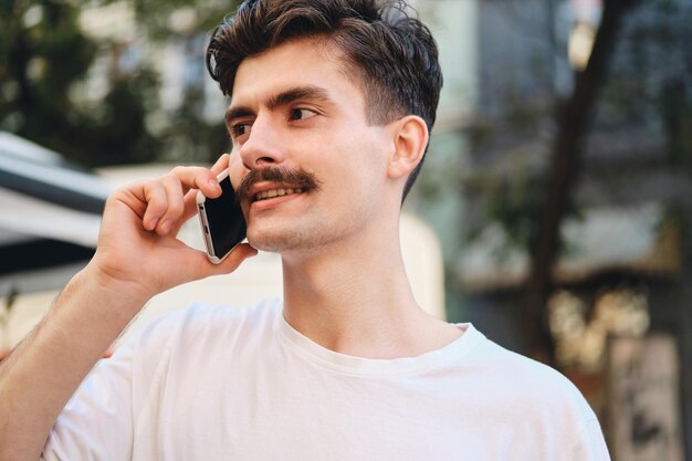 Ritratto di giovane uomo con i baffi belli in maglietta bianca che parla al cellulare mentre guarda sognante da parte sulla strada della città