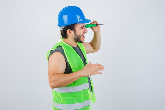 Ritratto di giovane uomo costruttore fingendo di sbirciare un buco con un cacciavite in uniforme e guardando vista frontale focalizzata