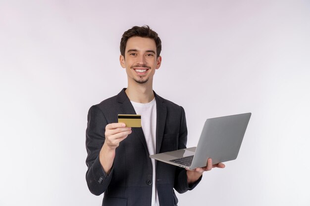 Ritratto di giovane uomo d'affari sorridente bello che tiene carta creadit e laptop in mano digitando e sfogliando pagine web isolate su sfondo bianco