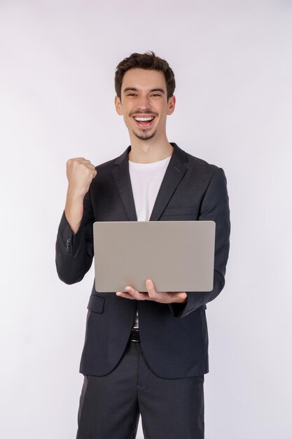 Ritratto di giovane uomo d'affari sorridente che tiene il laptop in mano digitando e navigando nelle pagine web mentre si fa un gesto vincente di pugno chiuso isolato su sfondo bianco