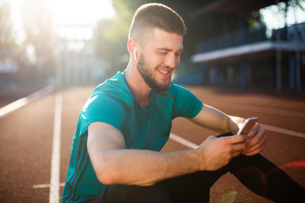 Ritratto di giovane uomo in auricolari wireless che usa gioiosamente il cellulare mentre trascorre del tempo sulla pista da corsa dello stadio