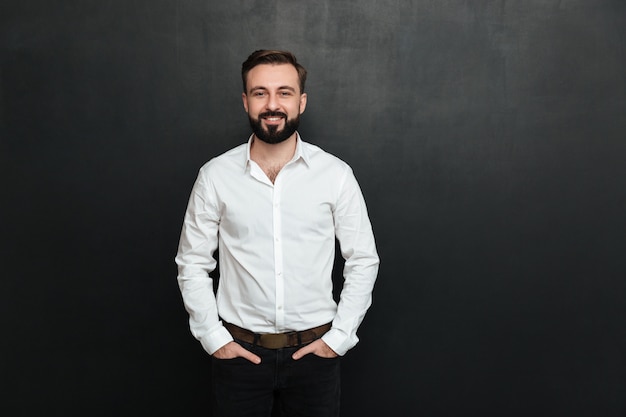 Ritratto di giovane uomo in camicia bianca in posa sulla fotocamera con ampio sorriso e le mani in tasca su grigio scuro