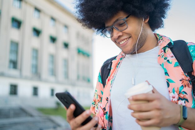 Ritratto di giovane uomo latino che usa il suo telefono cellulare e tiene una tazza di caffè mentre si cammina all'aperto per strada. Concetto urbano