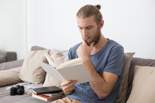 Ritratto di giovane uomo premuroso seduto sul grande divano grigio e libro di lettura a casa