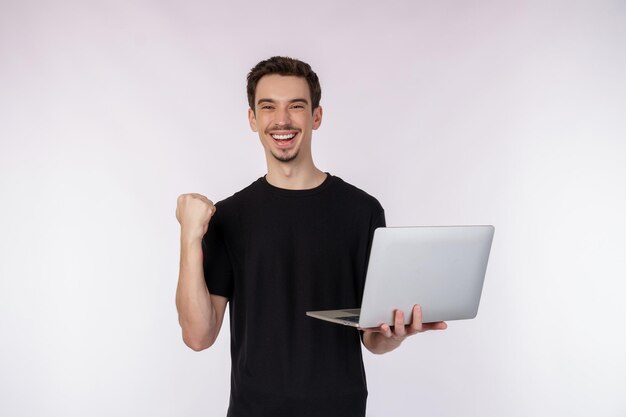 Ritratto di giovane uomo sorridente bello che tiene il laptop in mano digitando e navigando nelle pagine Web mentre si fa un gesto vincente di pugno chiuso isolato su sfondo bianco