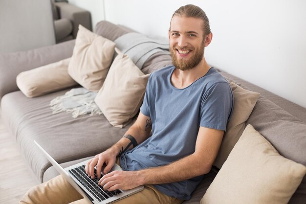 Ritratto di giovane uomo sorridente seduto su un grande divano grigio con laptop e felicemente guardando a porte chiuse