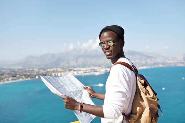 Ritratto di giovane viaggiatore afroamericano che guarda con la mappa cartacea tra le mani, indossa occhiali da sole e cappello, in piedi sulla piattaforma panoramica, ammirando la città europea e il bellissimo paesaggio marino