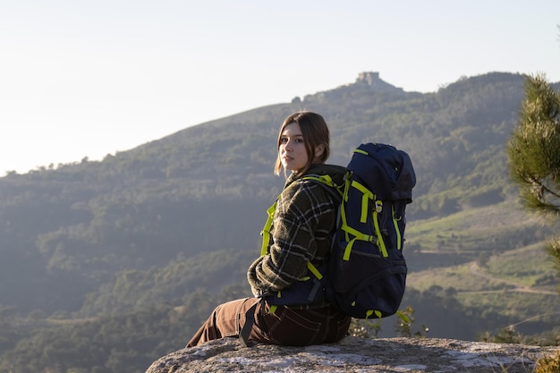 Ritratto di giovane viaggiatore con zaino e sacco a pelo femminile felice che si siede sulla pietra