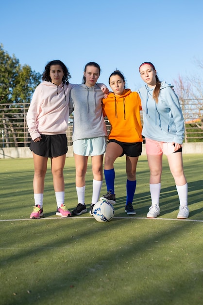 Ritratto di giovani calciatori femminili con palla sul campo. Squadra di quattro ragazze caucasiche che si preparano per l'allenamento mattutino abbracciando in piedi, guardando la fotocamera. Concetto di sport di squadra e stile di vita sano