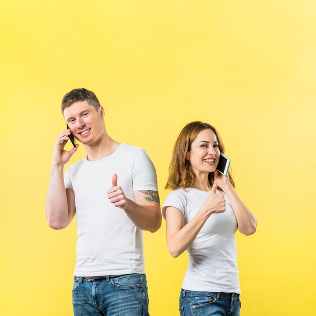 Ritratto di giovani coppie sorridenti che parlano sul telefono cellulare che mostra pollice sul segno contro il contesto giallo