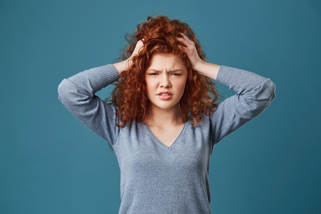 Ritratto di giovani womans di zenzero con capelli ondulati e le lentiggini che tengono le mani sulla testa stanca del mal di testa dopo una lunga giornata all'università.