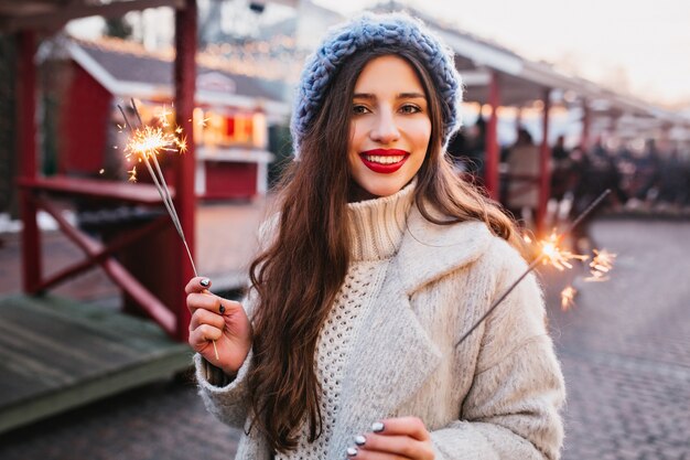 Ritratto di graziosa donna dai capelli castani in elegante camice bianco che tiene le luci del Bengala. Foto all'aperto di romantica ragazza europea in berretto blu in posa con stelle filanti sulla città di sfocatura