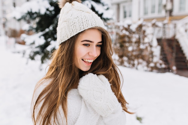 Ritratto di inverno accogliente di giovane donna gioiosa alla moda con capelli lunghi del brunette che cammina sulla strada piena di neve. Sorprese vere emozioni positive, guanti di lana bianca calda, cappello lavorato a maglia.