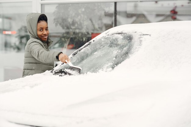 Ritratto di inverno della donna africana che pulisce la neve da un'auto. Donna in una giacca verde.