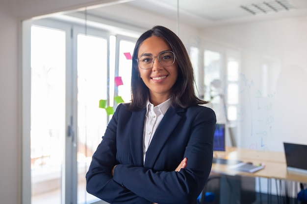 Ritratto di leader di affari latino sicuro. Giovane imprenditrice in tuta e occhiali in posa con le braccia conserte, guardando la fotocamera e sorridente. Concetto di leadership femminile